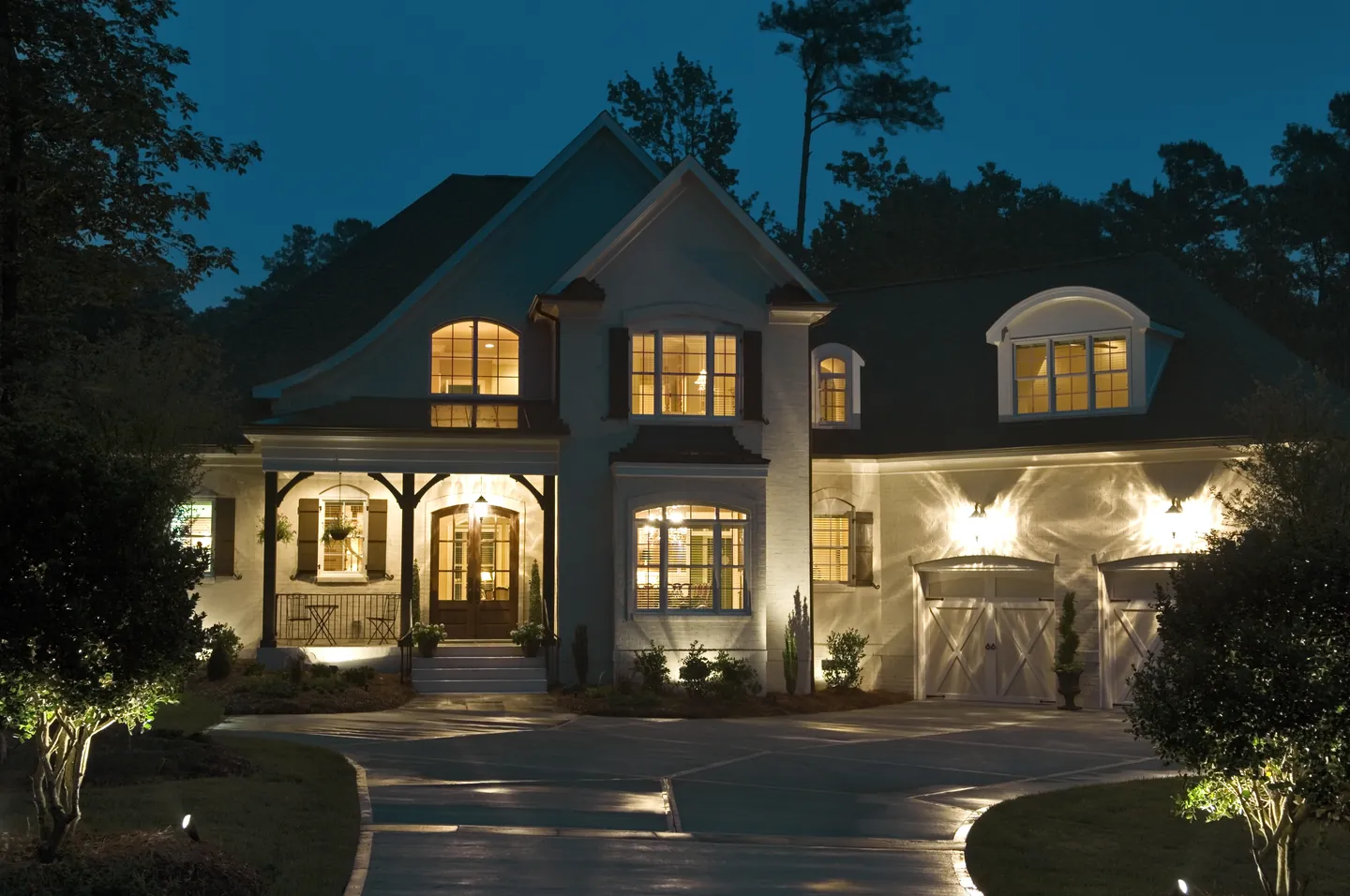 A cream gabled house at blue hour with its windows, porch, garage bays and the plantings along the drive all lit.