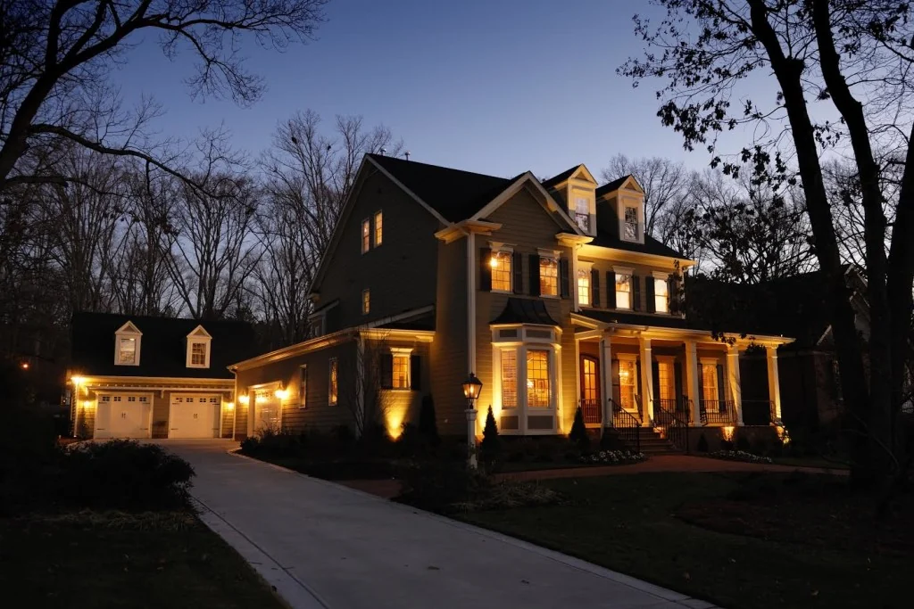 A colonial at dusk with its garage, porch, lantern post, bay window and the plantings along the walk all lit.