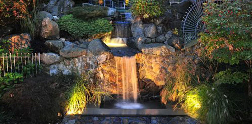 A garden waterfall over stone, lit from within the water at night.